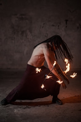 A person with long hair is performing with flaming staffs in a dimly lit environment, creating a dynamic and intense atmosphere.