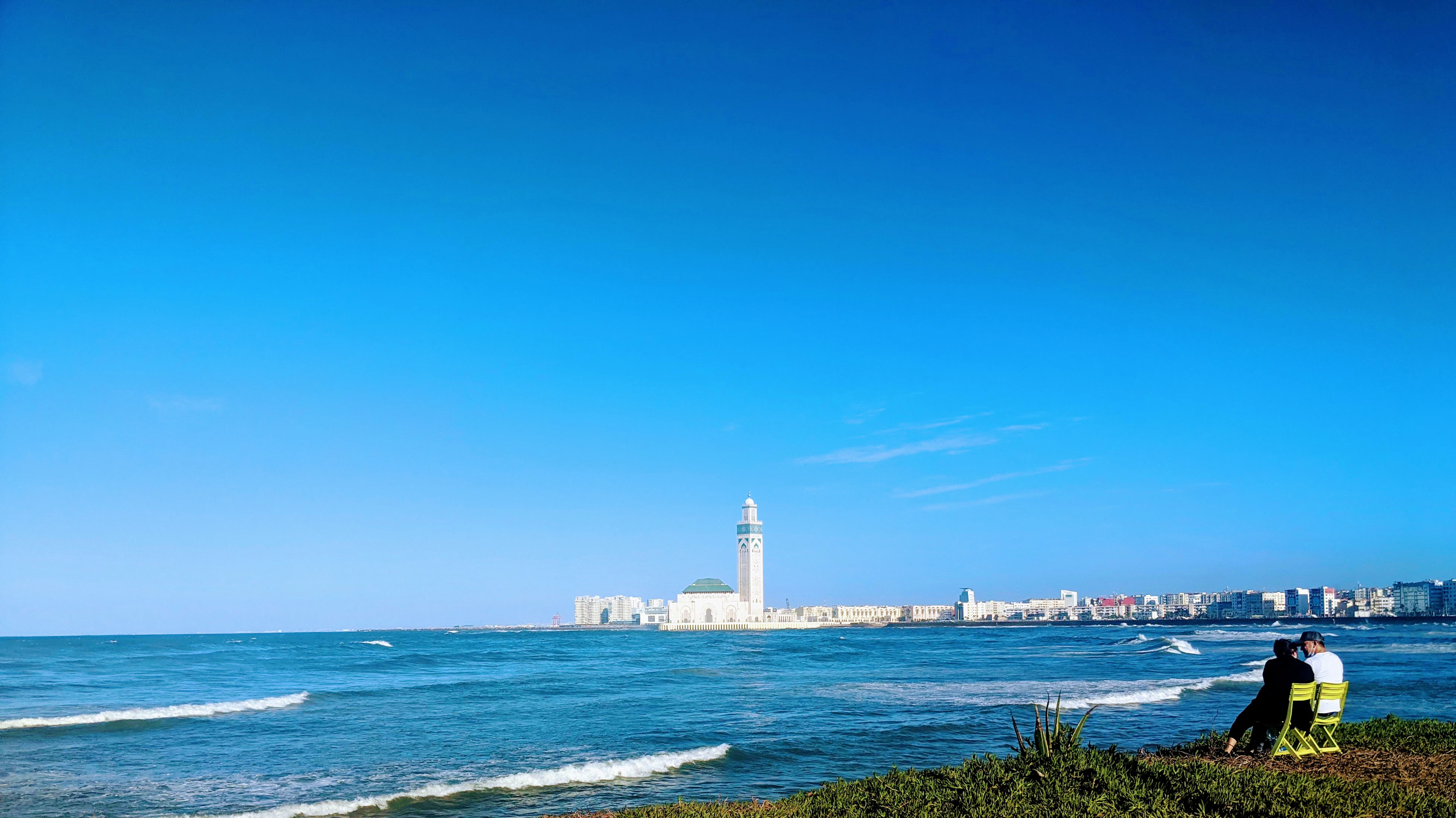 A couple sitting on the grass above the Atlantic surf on Casablanca's Corniche, with a tall white tower on the coastline in the distance
