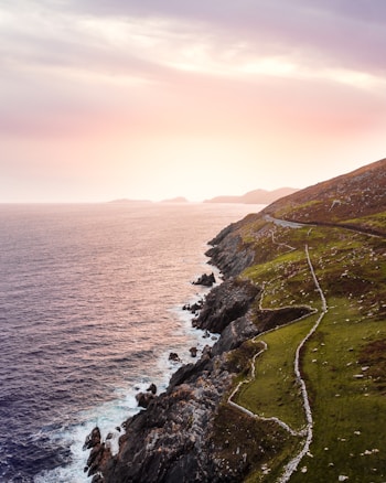 A picturesque coastal landscape with a winding path along the rugged cliffs. The ocean stretches out to the horizon, reflecting the soft colors of a sunset sky. The grassy hillside contrasts with the rocky shoreline, creating a serene and natural scene.