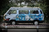 A white van with vibrant blue graffiti art featuring an abstract face and swirling designs. It is parked on a road beside dense greenery, with a traffic cone visible in the foreground.