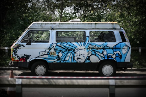 A white van with vibrant blue graffiti art featuring an abstract face and swirling designs. It is parked on a road beside dense greenery, with a traffic cone visible in the foreground.