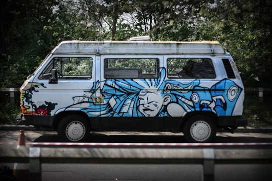 A white van with vibrant blue graffiti art featuring an abstract face and swirling designs. It is parked on a road beside dense greenery, with a traffic cone visible in the foreground.