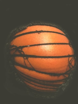 Close-up shot of a basketball partially wrapped in black thread. The orange texture of the ball is clearly visible against a dark background, with the threads creating a slightly chaotic pattern.
