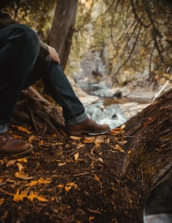 person in black pants and brown leather boots sitting on brown tree log during daytime