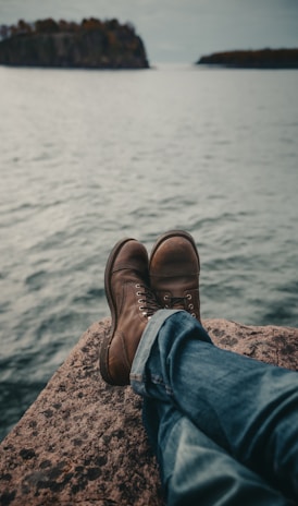 person in blue denim jeans and brown leather boots sitting on rock near body of water