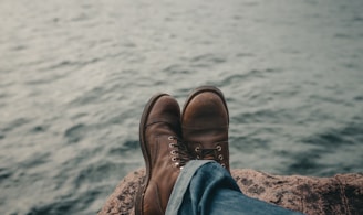 person in blue denim jeans and brown leather boots sitting on rock near body of water