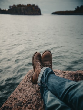 person in blue denim jeans and brown leather boots sitting on rock near body of water