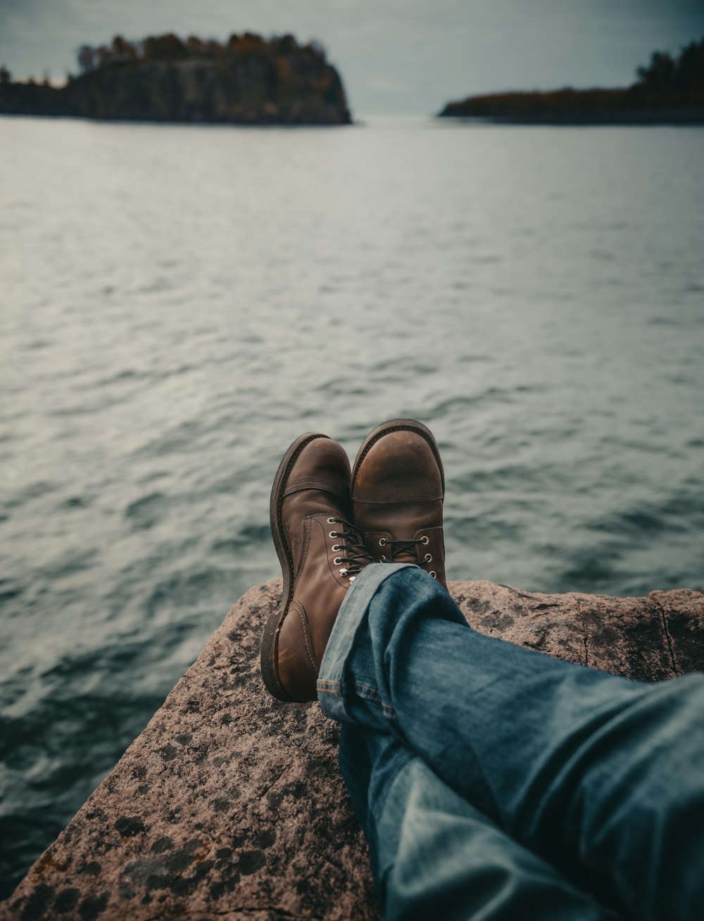 person in blue denim jeans and brown leather boots sitting on rock near body of water