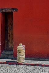 A tall stack of straw hats is placed on a cobblestone street in front of a vibrant red wall. The wooden door, slightly recessed, provides a rustic contrast to the bright backdrop.