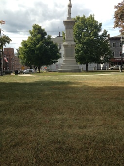 A public park features a prominent stone monument surrounded by well-maintained grass and lush, green trees. A backdrop of historic buildings and an overcast sky adds depth, while an American flag is faintly visible on the left side.