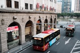 A city street scene with two buses on the road, one red and one green. The street is lined with a large, historic building featuring stone arches and a prominent sign in Chinese characters. Pedestrians are visible in the background near a river, and there are trees along the riverbank. A shop with bright red signage is situated on the left side under one of the arches.