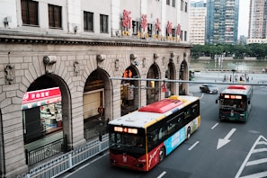 A city street scene with two buses on the road, one red and one green. The street is lined with a large, historic building featuring stone arches and a prominent sign in Chinese characters. Pedestrians are visible in the background near a river, and there are trees along the riverbank. A shop with bright red signage is situated on the left side under one of the arches.