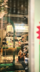 A bustling coffee shop interior with a transparent window reflection showing several people. Two baristas are working behind a counter displaying various beverages and menu signs. Customers are seated and appear to be engaged in conversation or enjoying their drinks. The floor features a patterned tile design, and the decor includes modern green and wooden elements.