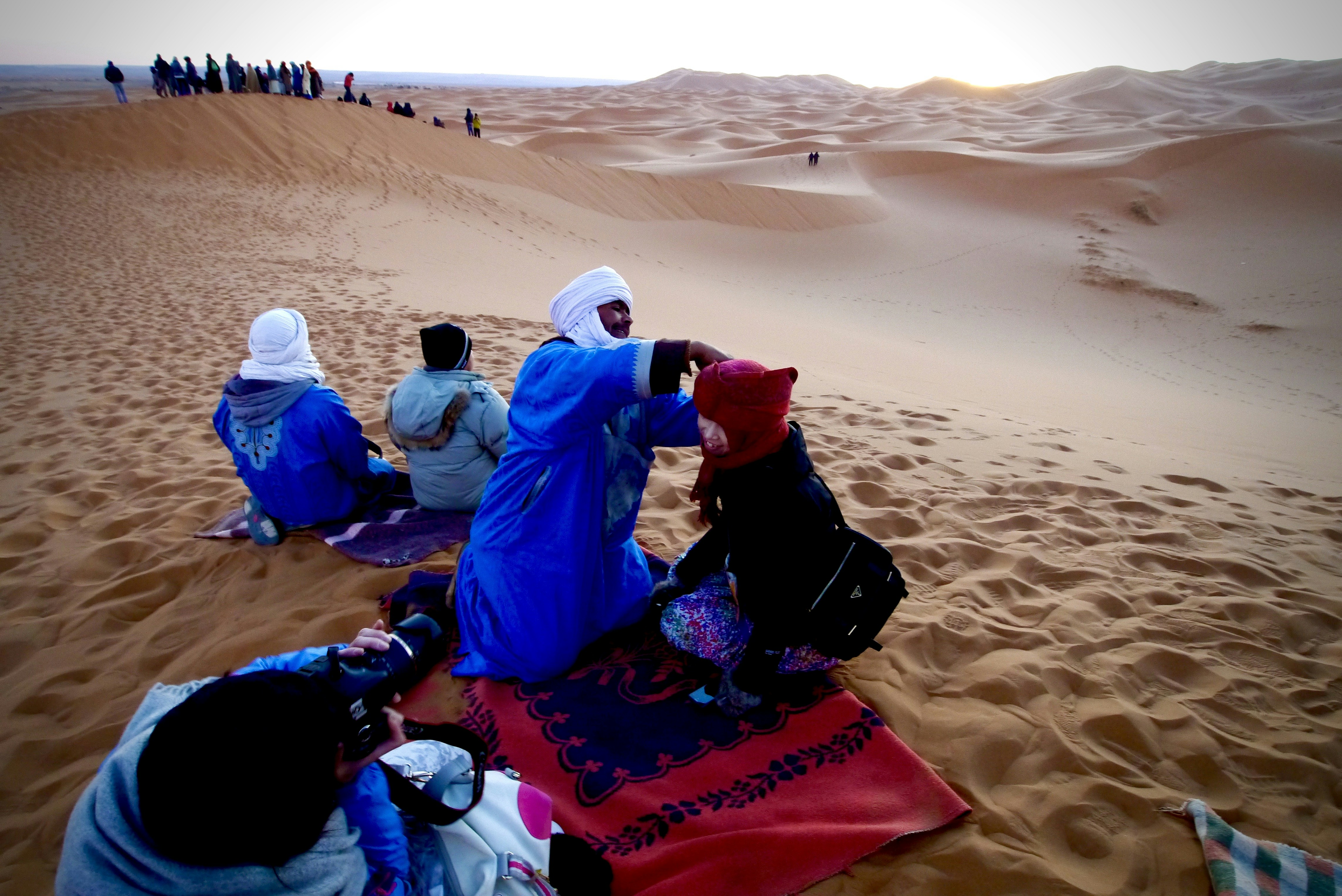 Man in white long sleeve shirt sitting on red textile on brown sand during daytime