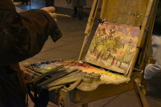 An outdoor scene features an artist painting on an easel, capturing a vibrant landscape that includes trees and figures. The artist's palette is laden with various colors, and brushes are scattered nearby. The setting appears to be dimly lit, possibly at dusk or at night.