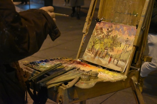 An outdoor scene features an artist painting on an easel, capturing a vibrant landscape that includes trees and figures. The artist's palette is laden with various colors, and brushes are scattered nearby. The setting appears to be dimly lit, possibly at dusk or at night.