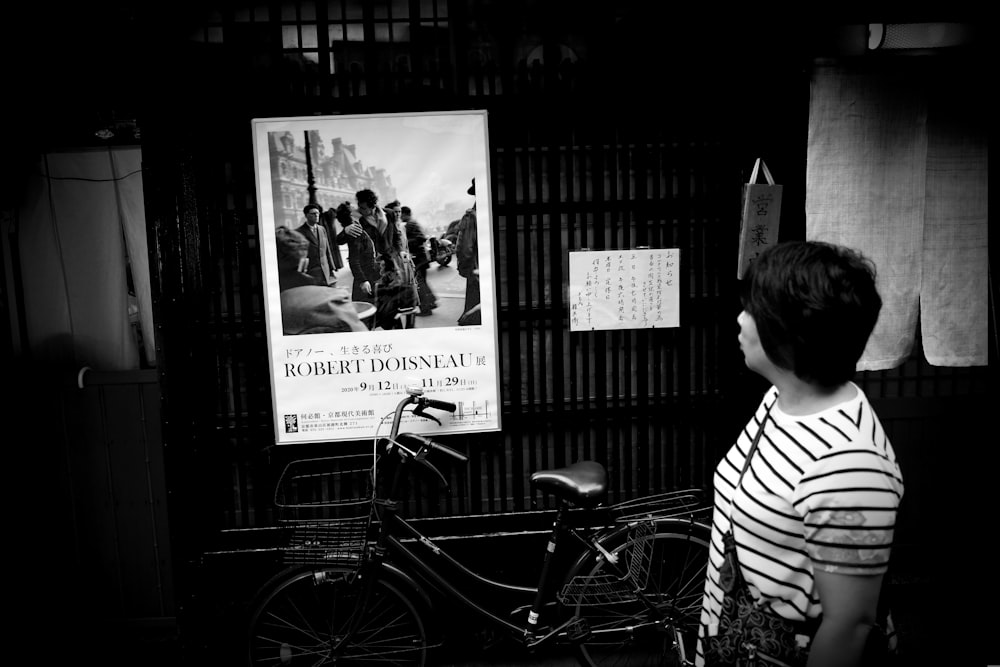 grayscale photo of woman in stripe shirt standing beside bicycle