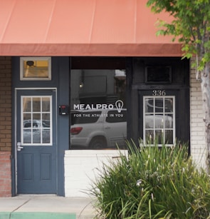 Front of a meal preparation business with a dark blue door and a large window displaying the name 'MEALPRO' and the slogan 'For the athlete in you.' The exterior features an orange awning and a small patch of greenery. A parked car is visible in the reflection of the window.