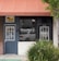 Front of a meal preparation business with a dark blue door and a large window displaying the name 'MEALPRO' and the slogan 'For the athlete in you.' The exterior features an orange awning and a small patch of greenery. A parked car is visible in the reflection of the window.