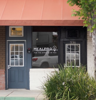 Front of a meal preparation business with a dark blue door and a large window displaying the name 'MEALPRO' and the slogan 'For the athlete in you.' The exterior features an orange awning and a small patch of greenery. A parked car is visible in the reflection of the window.