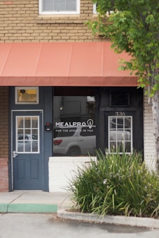 A storefront with a brick facade features a dark blue door and large window panels. The window displays the text 'MealPro For the Athlete in You.' An orange awning covers the entrance area, and there is a reflection of a car in the window. Green plants grow along the sidewalk edge, adding a natural element to the urban scene. The building number 336 is visible above the door.