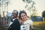 A bride and groom couple smiling joyfully, standing together in a flower-filled meadow.