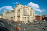 A large, historic-looking hotel building with a classic architectural style is situated in a busy urban area. The street surrounding the hotel is bustling with activity, including pedestrians and various vehicles. The sky is blue with some scattered clouds adding to the vibrancy of the scene.