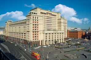 A large, historic-looking hotel building with a classic architectural style is situated in a busy urban area. The street surrounding the hotel is bustling with activity, including pedestrians and various vehicles. The sky is blue with some scattered clouds adding to the vibrancy of the scene.