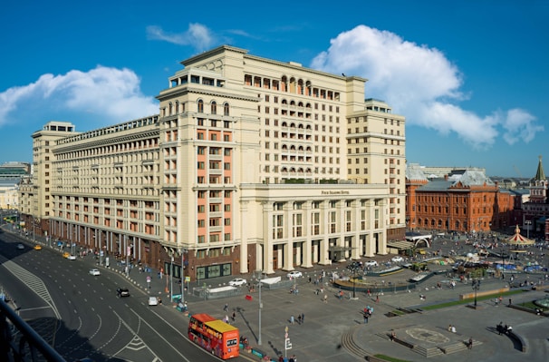 A large, historic-looking hotel building with a classic architectural style is situated in a busy urban area. The street surrounding the hotel is bustling with activity, including pedestrians and various vehicles. The sky is blue with some scattered clouds adding to the vibrancy of the scene.