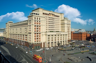 A large, historic-looking hotel building with a classic architectural style is situated in a busy urban area. The street surrounding the hotel is bustling with activity, including pedestrians and various vehicles. The sky is blue with some scattered clouds adding to the vibrancy of the scene.
