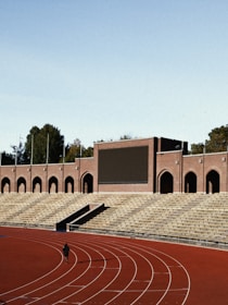 An empty stadium with a large track and field area. The seating is constructed with tiered rows of stone bleachers. A brick structure with arched doorways and a large blank screen dominates the background. A single person is seen walking or running on the track, which is red and lined with white lanes. Trees are visible in the background behind the stadium.