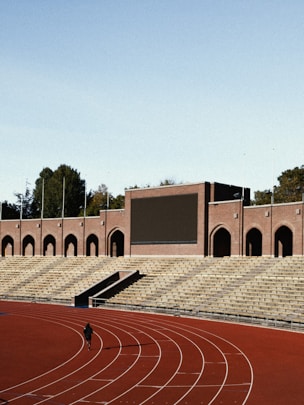 An empty stadium with a large track and field area. The seating is constructed with tiered rows of stone bleachers. A brick structure with arched doorways and a large blank screen dominates the background. A single person is seen walking or running on the track, which is red and lined with white lanes. Trees are visible in the background behind the stadium.