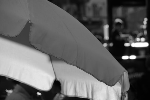 A large fabric umbrella with visible stitching and textured material dominates the foreground. In the background, there are blurred lights and shadowy figures, suggesting an outdoor or cafe setting with bokeh effect.