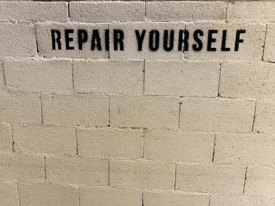 A concrete brick wall features the stencil-painted message 'REPAIR YOURSELF' in bold black letters.