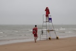 Two lifeguards are present on a sandy beach with grey overcast skies. One lifeguard stands on an elevated platform, looking through binoculars towards the sea, while the other approaches with a rescue backpack. The ocean is calm with small waves, and there is a sign with logos on the lifeguard stand.