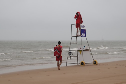 Two lifeguards are present on a sandy beach with grey overcast skies. One lifeguard stands on an elevated platform, looking through binoculars towards the sea, while the other approaches with a rescue backpack. The ocean is calm with small waves, and there is a sign with logos on the lifeguard stand.