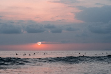 A serene ocean scene at sunset with gentle waves rolling in. Silhouettes of people can be seen in the water, creating a peaceful and communal atmosphere. The sky is painted with subtle hues of pink, orange, and gray.