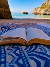 An open book lies on a patterned blue and white fabric, set on a sandy beach with a clear view of the sea and rocky cliffs in the background. The scene combines the elements of nature and relaxation.