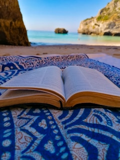 An open book lies on a patterned blue and white fabric, set on a sandy beach with a clear view of the sea and rocky cliffs in the background. The scene combines the elements of nature and relaxation.