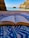 An open book lies on a patterned blue and white fabric, set on a sandy beach with a clear view of the sea and rocky cliffs in the background. The scene combines the elements of nature and relaxation.