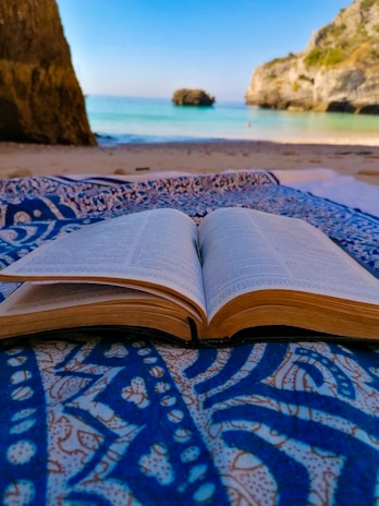 An open book lies on a patterned blue and white fabric, set on a sandy beach with a clear view of the sea and rocky cliffs in the background. The scene combines the elements of nature and relaxation.