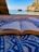 An open book lies on a patterned blue and white fabric, set on a sandy beach with a clear view of the sea and rocky cliffs in the background. The scene combines the elements of nature and relaxation.