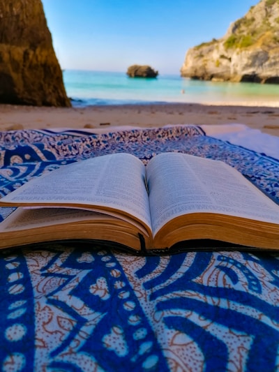 An open book lies on a patterned blue and white fabric, set on a sandy beach with a clear view of the sea and rocky cliffs in the background. The scene combines the elements of nature and relaxation.