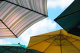 Four large outdoor umbrellas with different colors and patterns are opened against a clear blue sky. The umbrellas are positioned at various angles, and their shapes create a geometric pattern. The colors include striped patterns, green, and yellow.