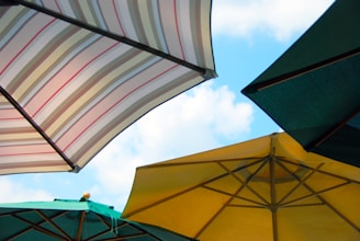 Four large outdoor umbrellas with different colors and patterns are opened against a clear blue sky. The umbrellas are positioned at various angles, and their shapes create a geometric pattern. The colors include striped patterns, green, and yellow.