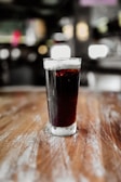 A tall glass filled with a dark, fizzy beverage, possibly cola, sits on a wooden surface. The glass has condensation on its exterior, indicating a cold temperature. The background is blurred, creating a bokeh effect with lights and indistinguishable shapes.