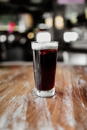 A tall glass filled with a dark, fizzy beverage, possibly cola, sits on a wooden surface. The glass has condensation on its exterior, indicating a cold temperature. The background is blurred, creating a bokeh effect with lights and indistinguishable shapes.