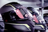 A close-up view of several high-tech motorcycle helmets arranged in a row. The helmets feature a sleek, aerodynamic design with a combination of glossy black and reflective yellow patterns. The visors have warning stickers indicating anti-scratch features, with text and symbols visible upon inspection.