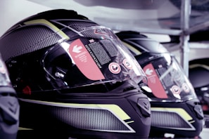 A close-up view of several high-tech motorcycle helmets arranged in a row. The helmets feature a sleek, aerodynamic design with a combination of glossy black and reflective yellow patterns. The visors have warning stickers indicating anti-scratch features, with text and symbols visible upon inspection.