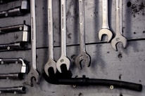 A collection of open-end and box-end wrenches hang on a weathered wooden board. The metal tools are neatly organized in a row, displaying their shiny metallic surfaces. A black hose or pipe with fittings is also visible below the wrenches.